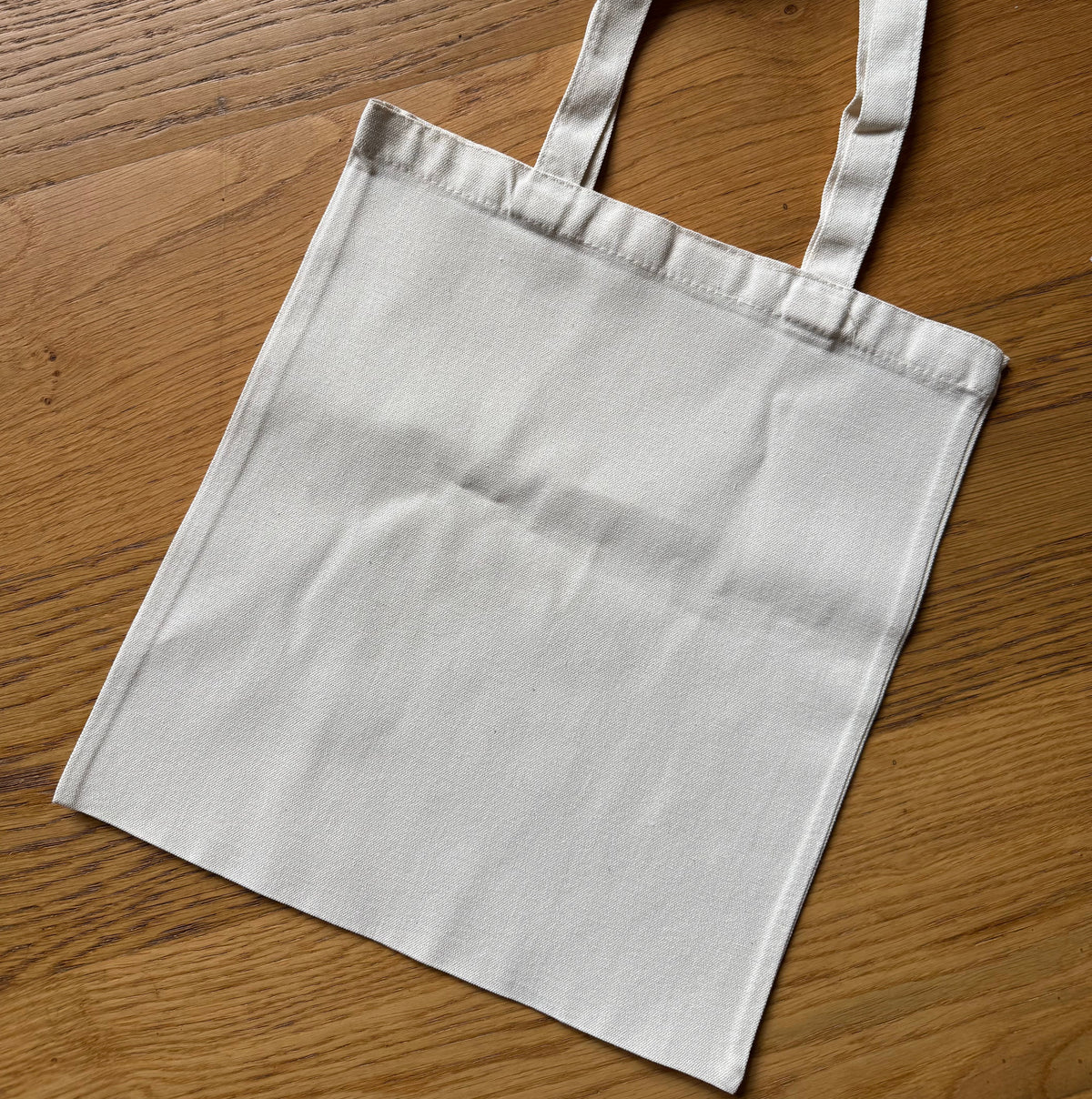 A plain canvas tote bag in a light off-white color, laid flat on a warm brown wooden surface.