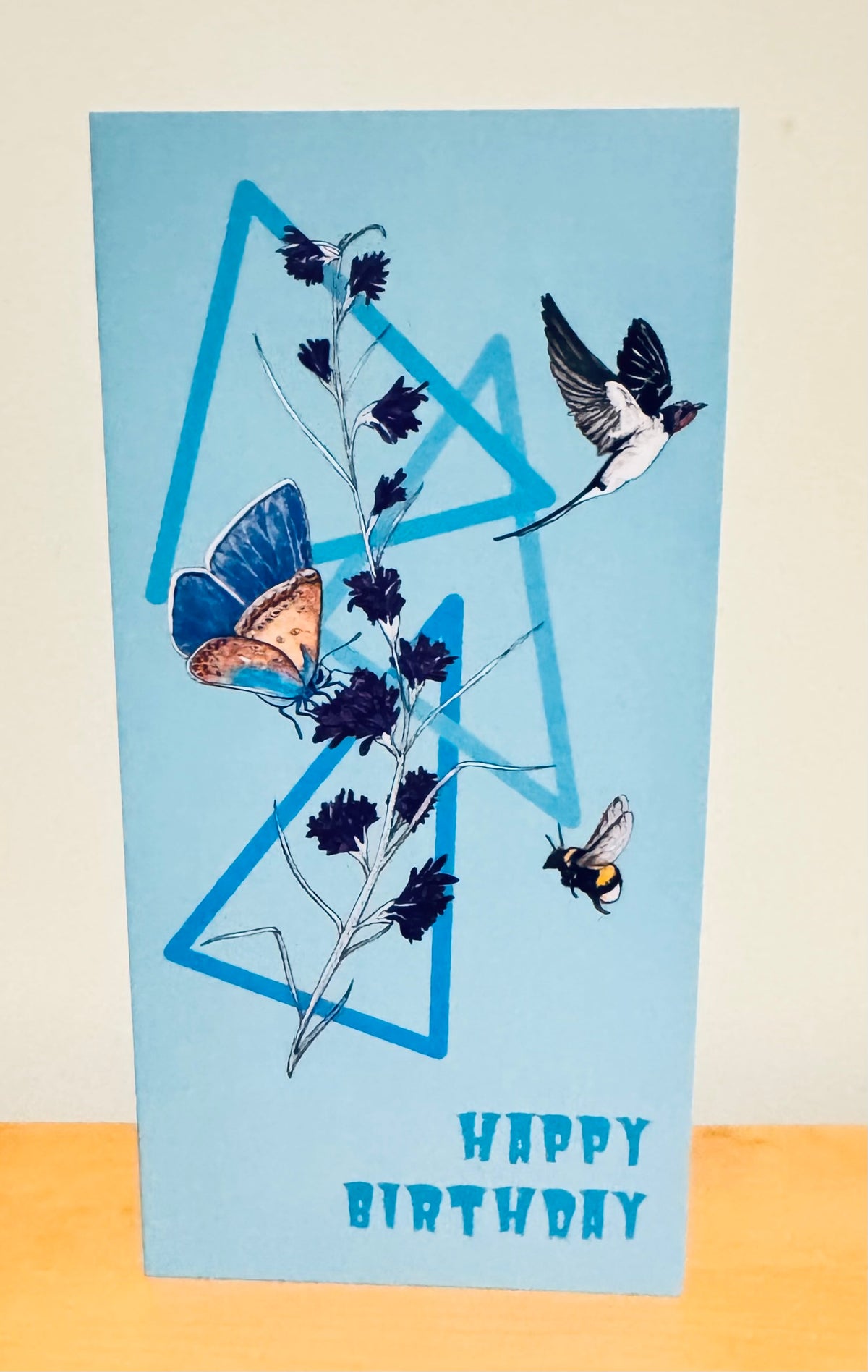 A light blue greeting card with a 'Happy Birthday' design, featuring blue geometric triangles, a butterfly, a bird, and a bee on a floral stem, standing on a wooden surface against a neutral background.