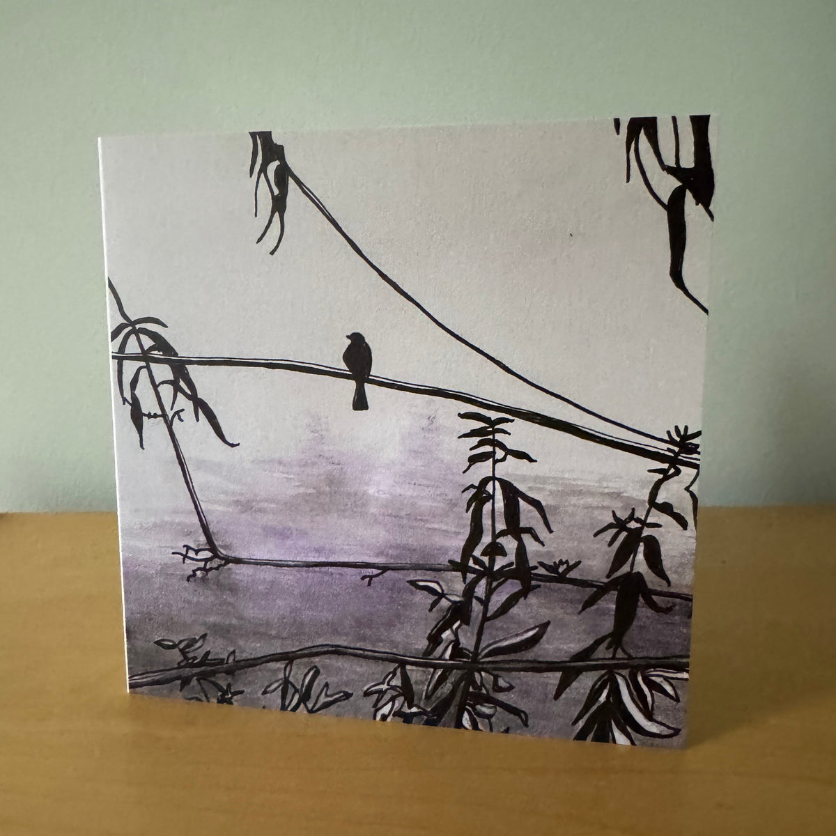 A square art print or card featuring a minimalist black ink illustration of a bird perched on branches against a pale lavender and gray background, standing on a wooden surface against a light green wall.