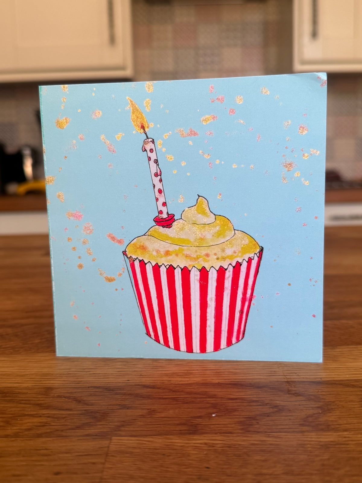 A square greeting card featuring a hand-drawn illustration of a cupcake with red and white striped paper liner, yellow frosting, and a single candle on a light blue background, standing on a wooden table.