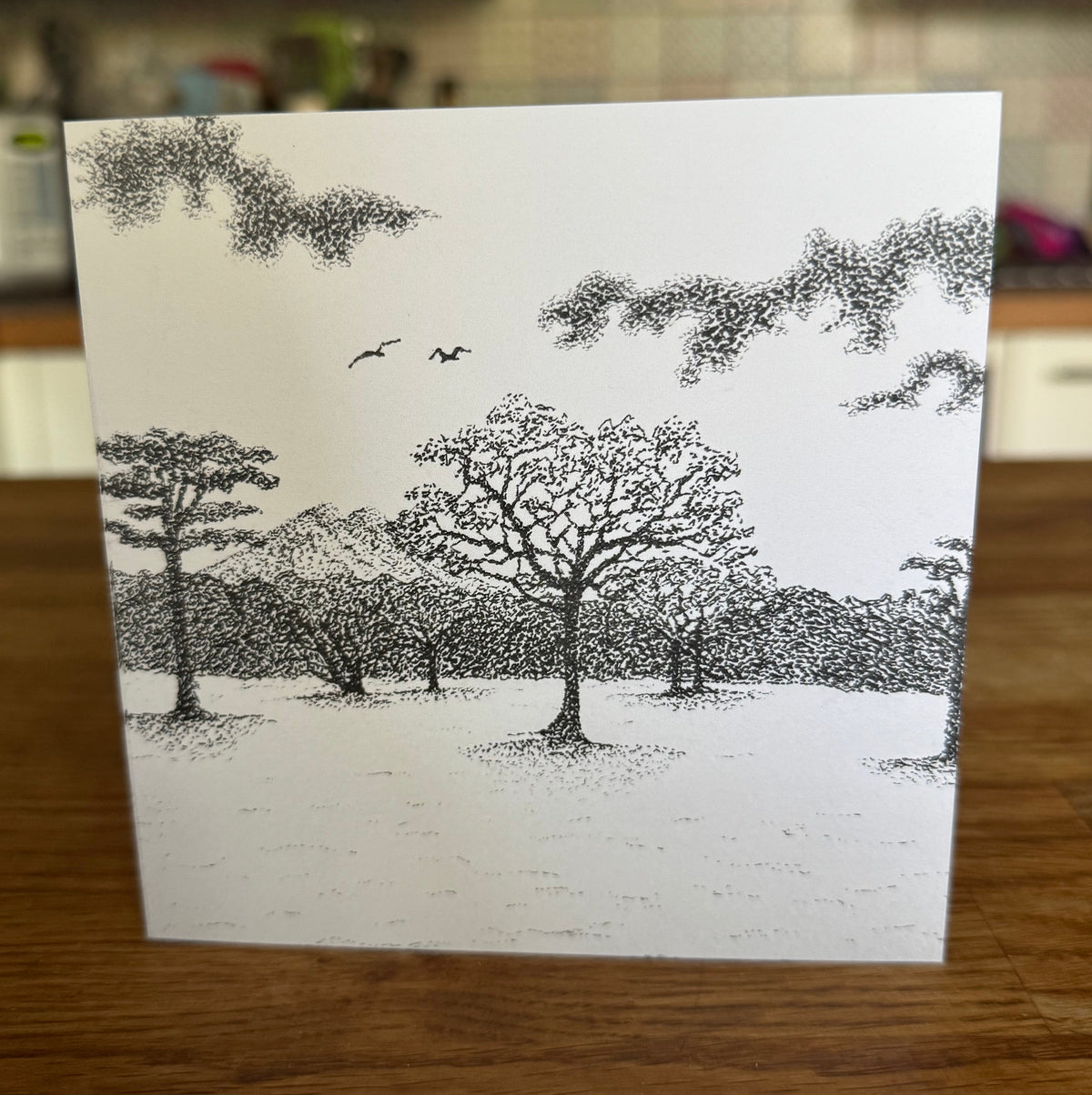 A square white art print or card featuring a black ink-style illustration of trees, birds, and mountains, standing on a wooden table with a blurred kitchen in the background.