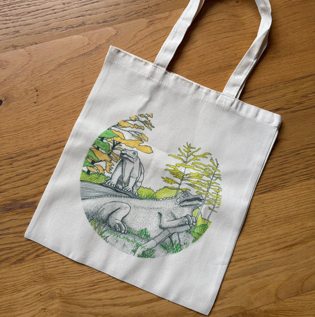 A white canvas tote bag with long handles, featuring a circular printed illustration of dinosaurs in a prehistoric landscape, displayed on a wooden surface.