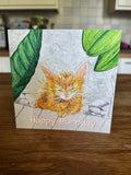 A square greeting card featuring a colored pencil drawing of an orange tabby kitten under green leaves, with the text 'Happy Purrday' at the bottom, displayed on a wooden table in a kitchen setting.