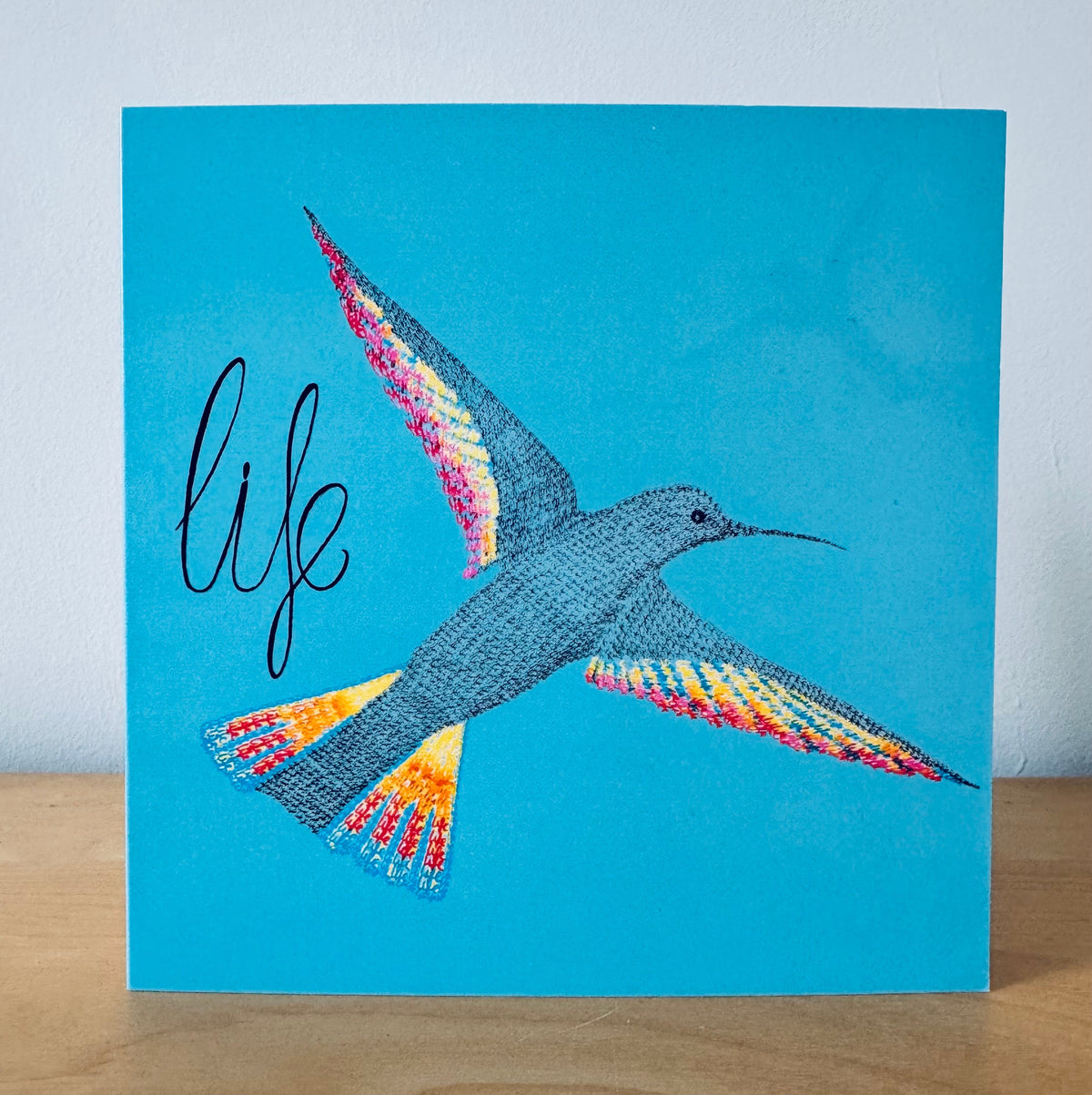 A square art print or card featuring a stylized hummingbird in flight on a bright turquoise background, with the word "life" in black script on the left. The print is photographed standing on a wooden surface against a light wall.