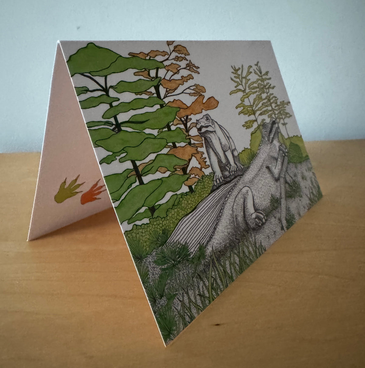 A folded art print or greeting card standing on a wooden surface against a light blue-gray wall. The front shows a detailed black-and-white illustration of a dinosaur in a grassy landscape with green and brown trees, while the inside flap shows a small orange and green footprint.
