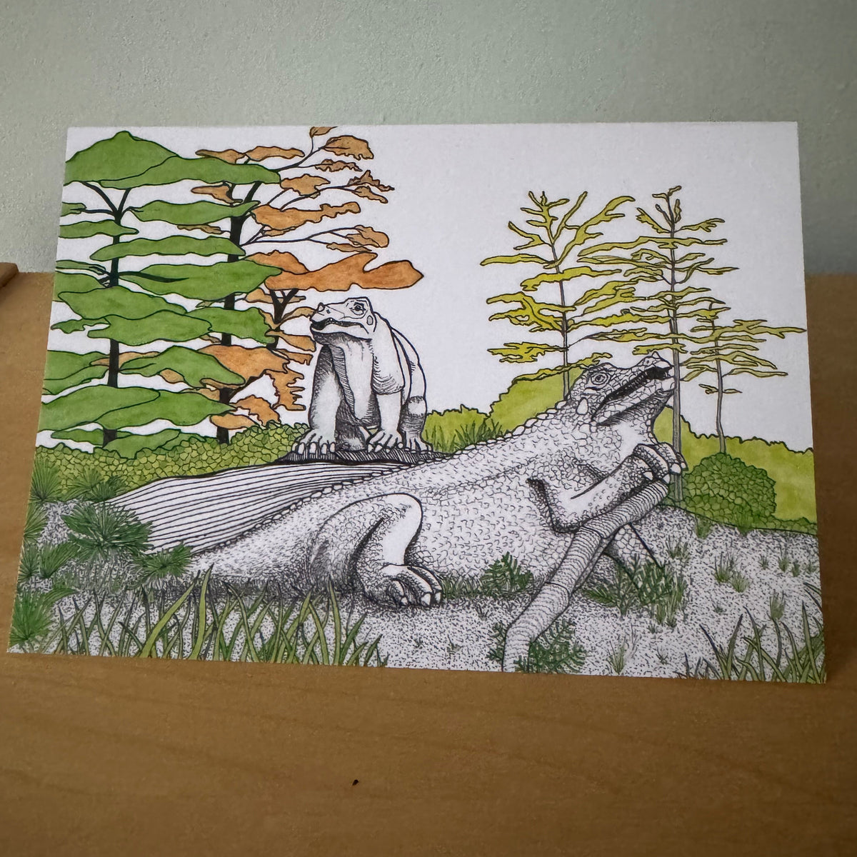 A rectangular art card featuring a black-and-white sketch of two iguanodon dinosaurs with green and orange trees in the background, displayed on a wooden surface against a light gray wall.