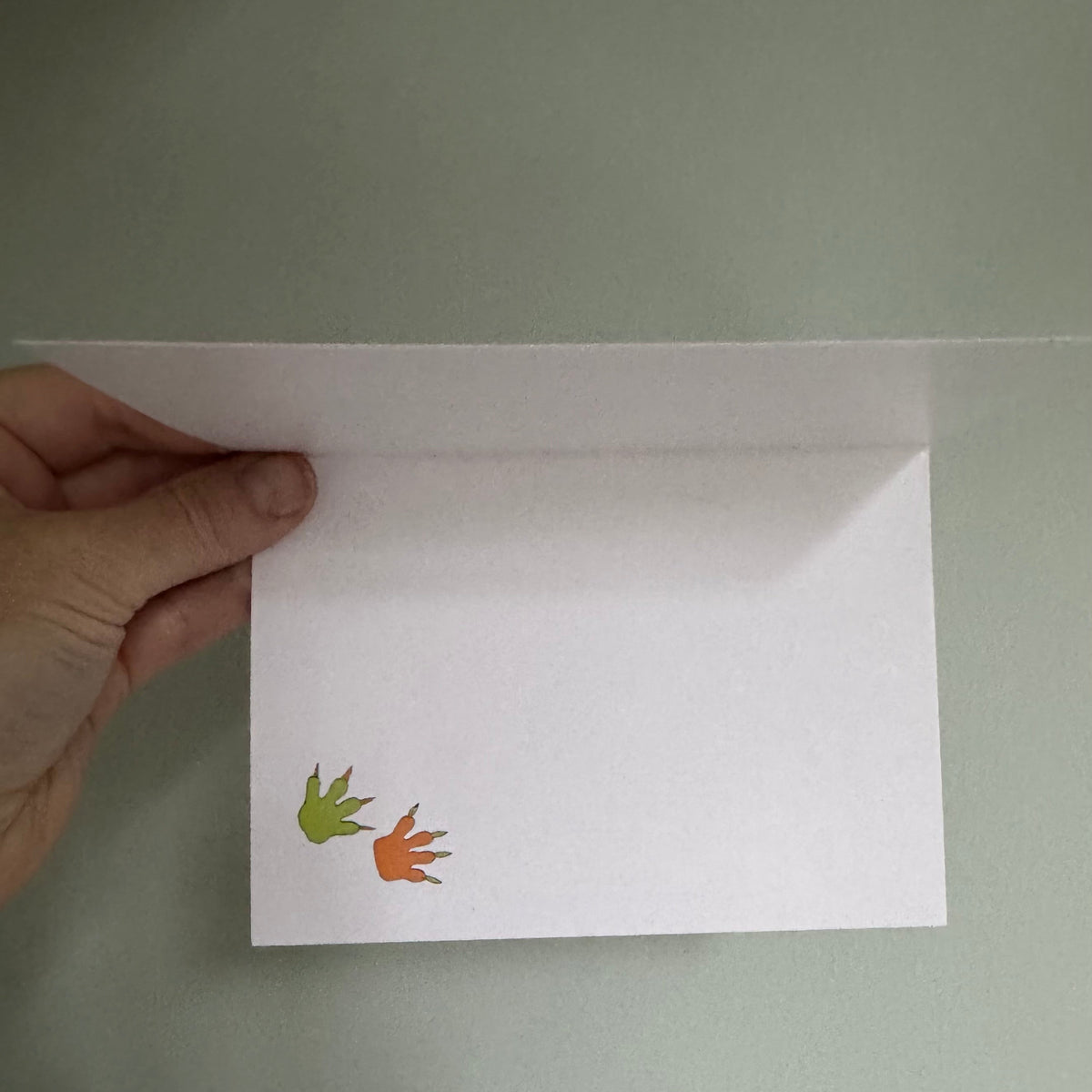 A hand holds a white square card against a muted green-gray background. The card has a small green footprint and an orange footprint printed near the bottom left corner.