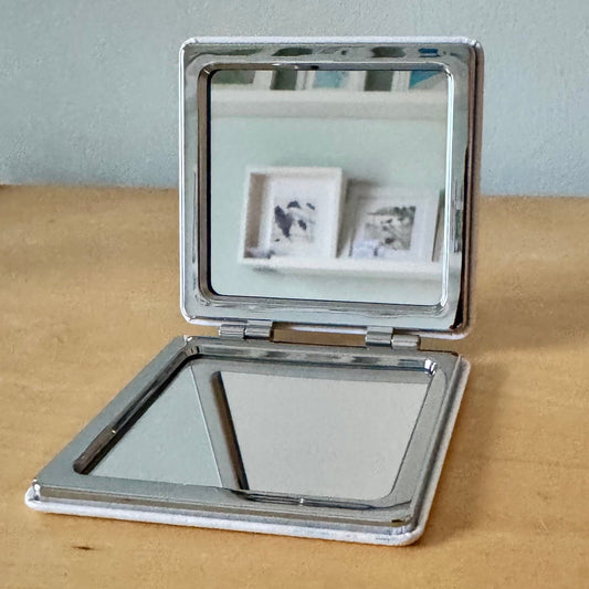 A square compact mirror with a polished silver metal frame, shown open on a light wood tabletop against a pale blue-gray wall. The top half reflects two small framed pictures, and the bottom half shows a plain mirror surface.
