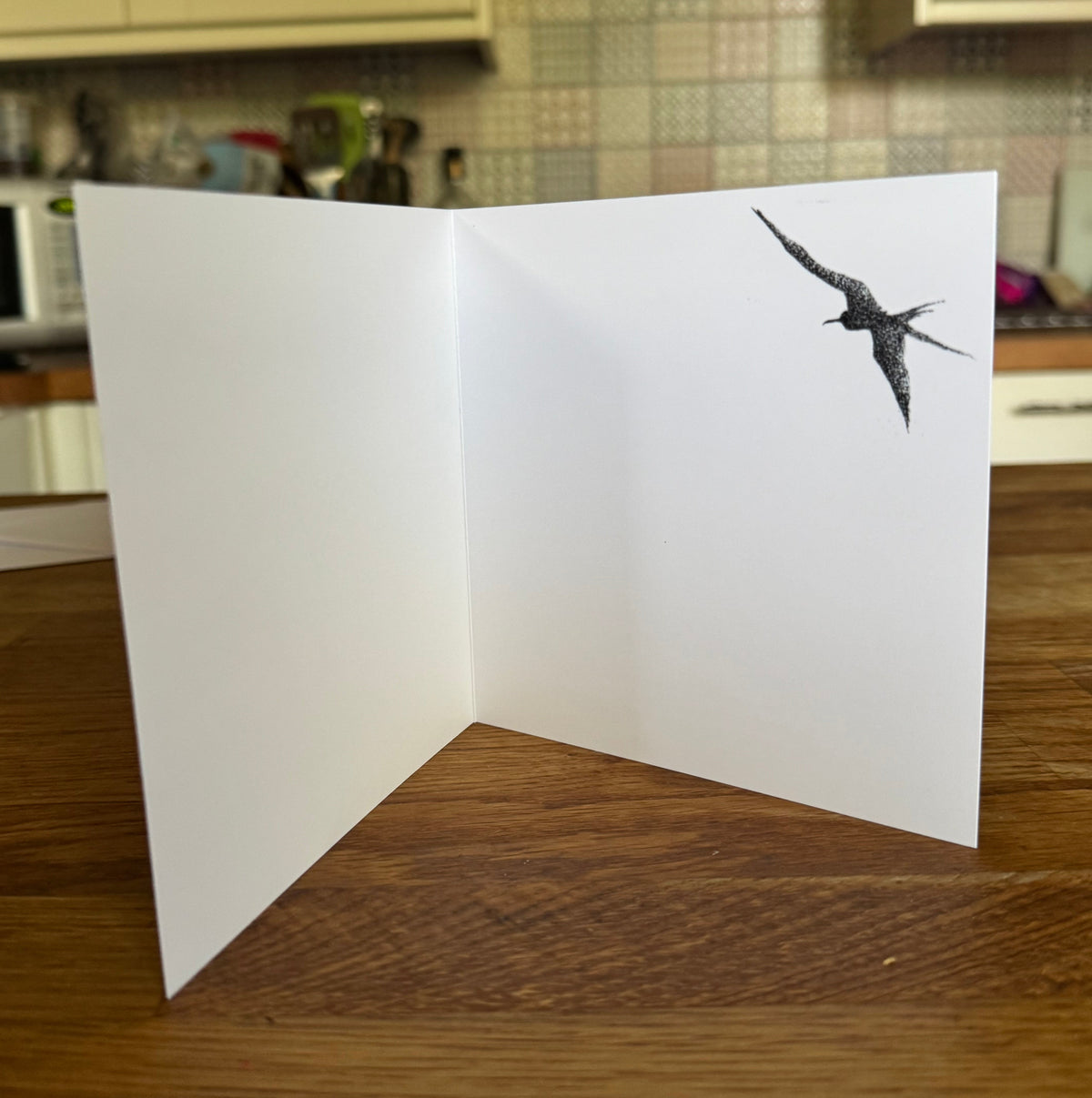 A white open greeting card standing on a wooden table, featuring a black ink illustration of a flying bird on the right panel, with a blurred kitchen background.