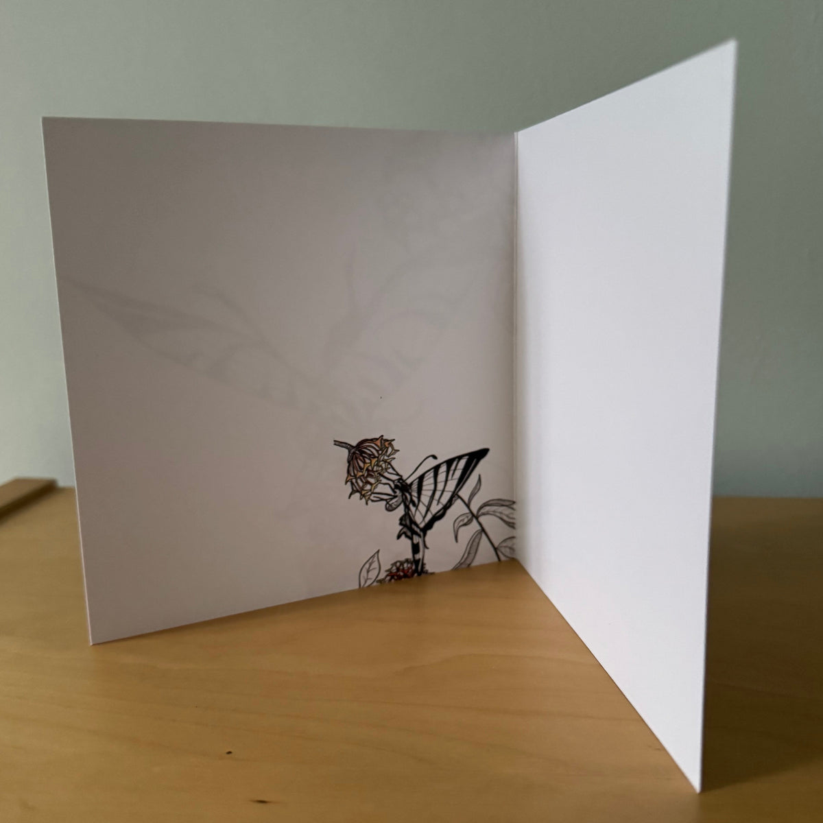 A white folded greeting card standing open on a light wooden table against a pale green wall. The left panel features a delicate black line drawing of a bird perched on a branch.