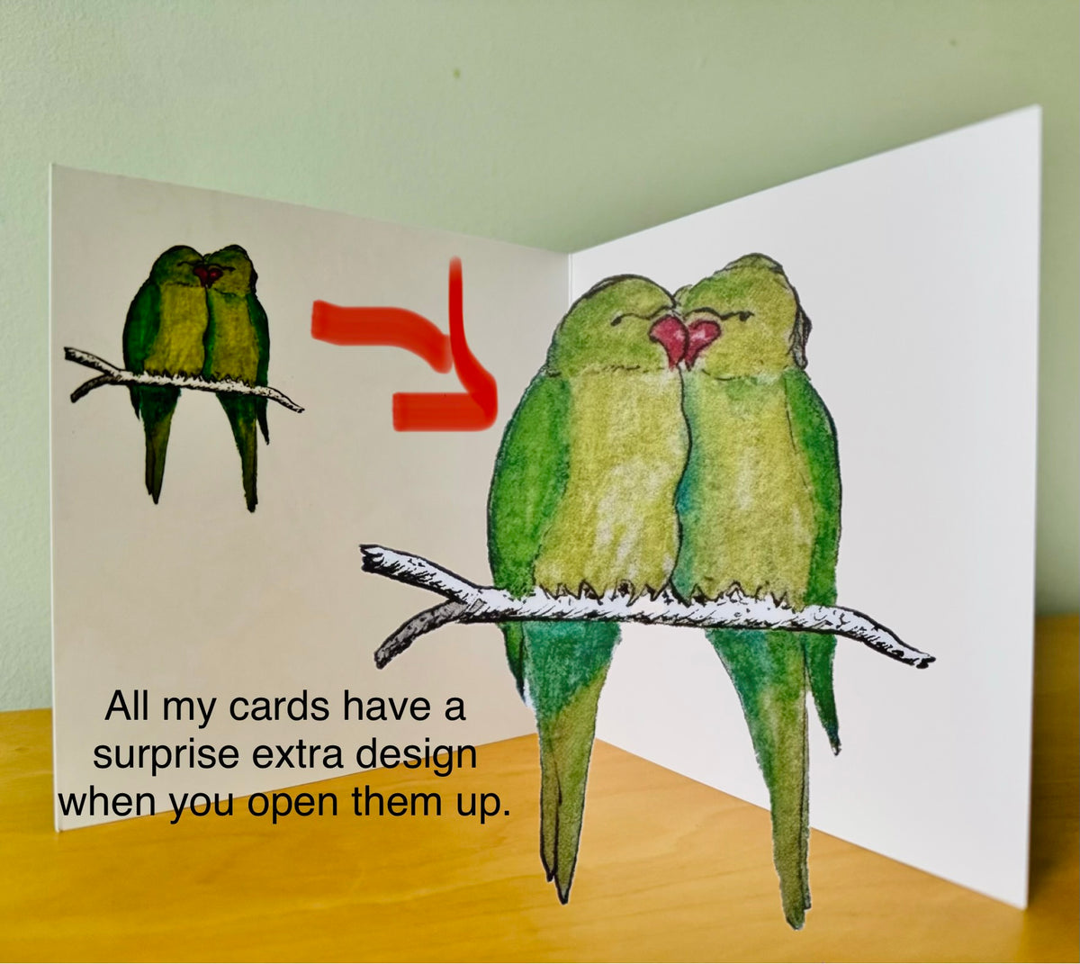 An open greeting card displayed on a wooden surface against a pale green wall. The card features a watercolor illustration of two green birds on a branch, with a red arrow pointing to a larger version of the same design on the inside panel.
