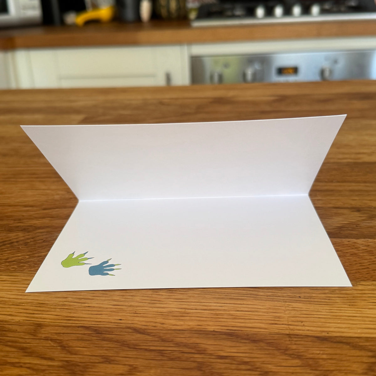 A white folded greeting card with two small colourful footprints in green and blue, placed on a wooden table with a blurred kitchen background.