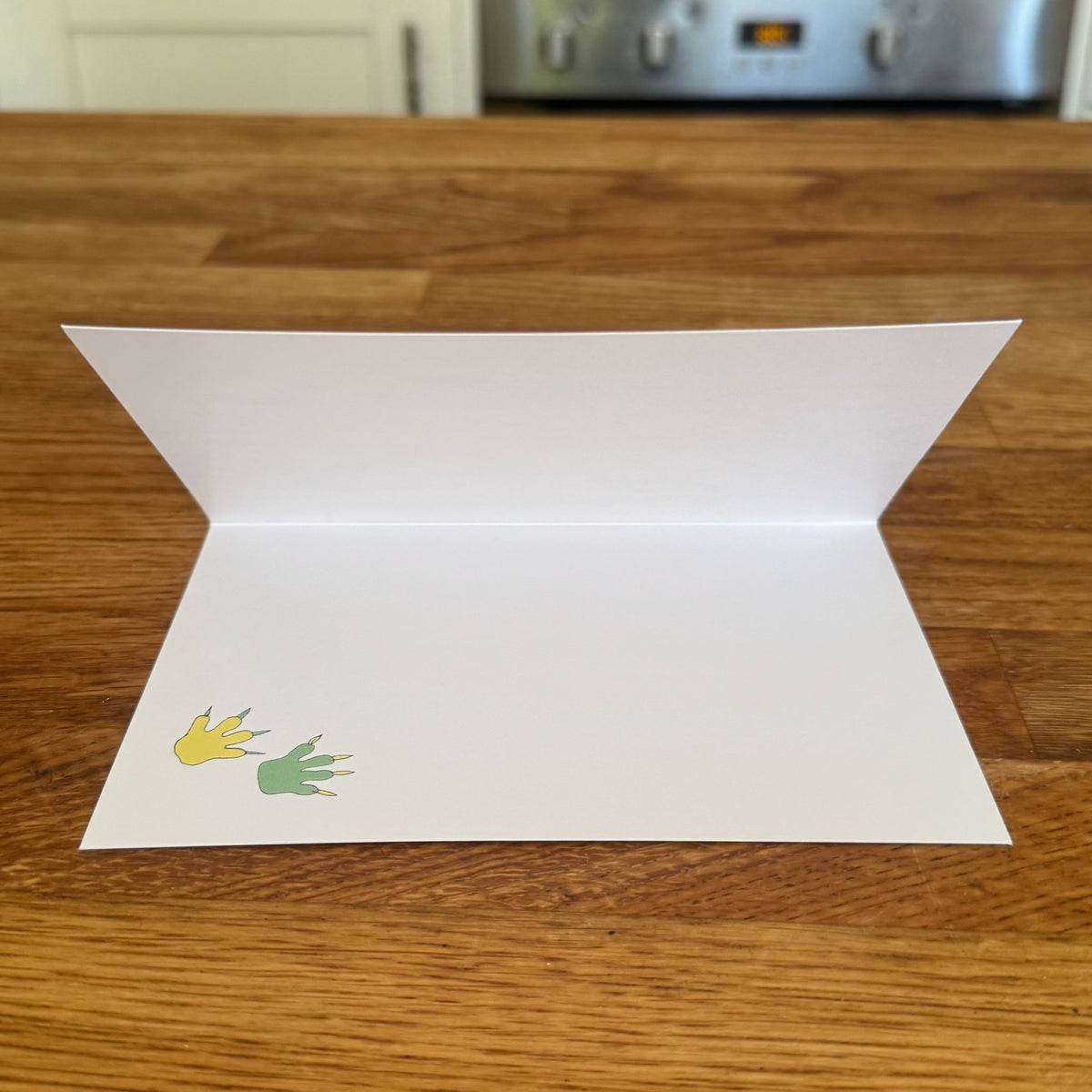 A white folded greeting card or paper plate-style card with two small footprints in yellow and green, displayed open on a wooden kitchen countertop with a blurred stove and cabinets in the background.