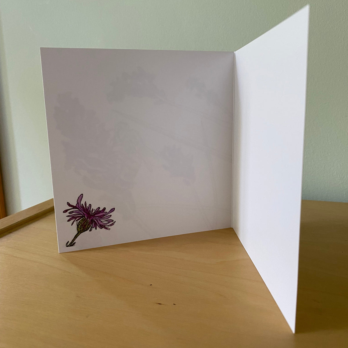 A white folded greeting card standing open on a light wood tabletop, with a small purple flower illustration in the lower left corner of the left panel and a pale green background behind it.