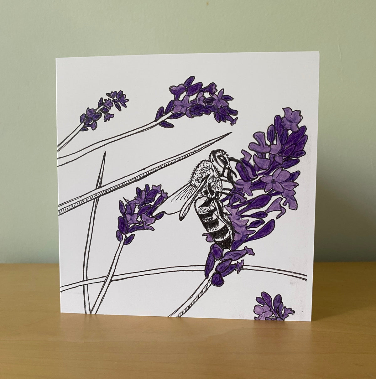 A square white art print or card featuring a black-and-purple illustration of a bee on lavender flowers, standing on a wooden surface against a pale green wall.
