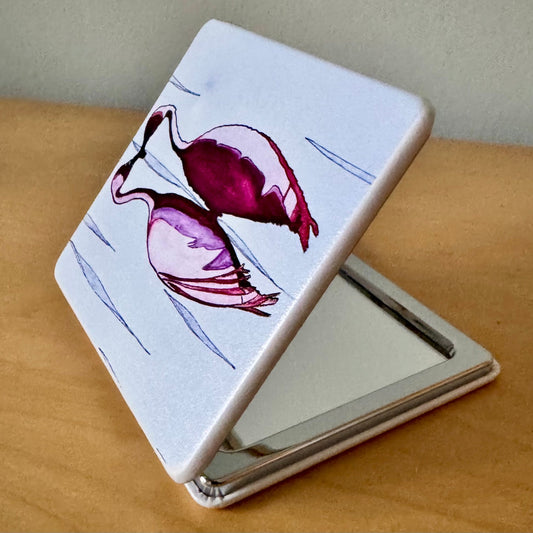 A square compact mirror with a white cover featuring a painted pink flamingo design, shown open on a wooden surface.