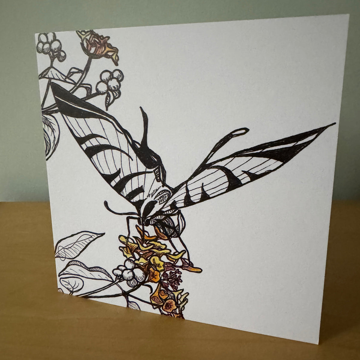 A square white art print or card featuring a black line-drawn butterfly perched on a floral branch with small warm-toned colored flowers, photographed standing on a wooden surface against a pale green wall.