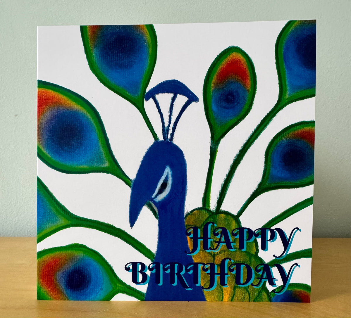 A square birthday card featuring a painted peacock with blue, green, and orange feather details and the words 'HAPPY BIRTHDAY' in blue script, standing on a wooden surface against a light wall.