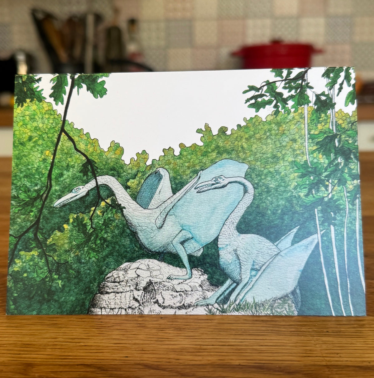 A rectangular card featuring an illustration of two light blue Crystal Palace pterosaurs among green trees, displayed on a wooden table with a blurred kitchen background.