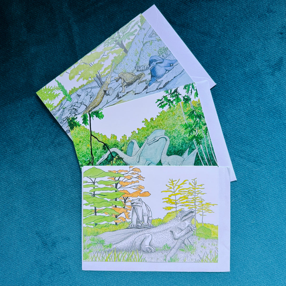 Three illustrated cards or art prints fanned out on a textured teal-blue background. The cards feature hand-drawn and painted prehistoric and forest scenes, including a dinosaurs and trees, with one card showing black-and-white line art of a dinosaur.