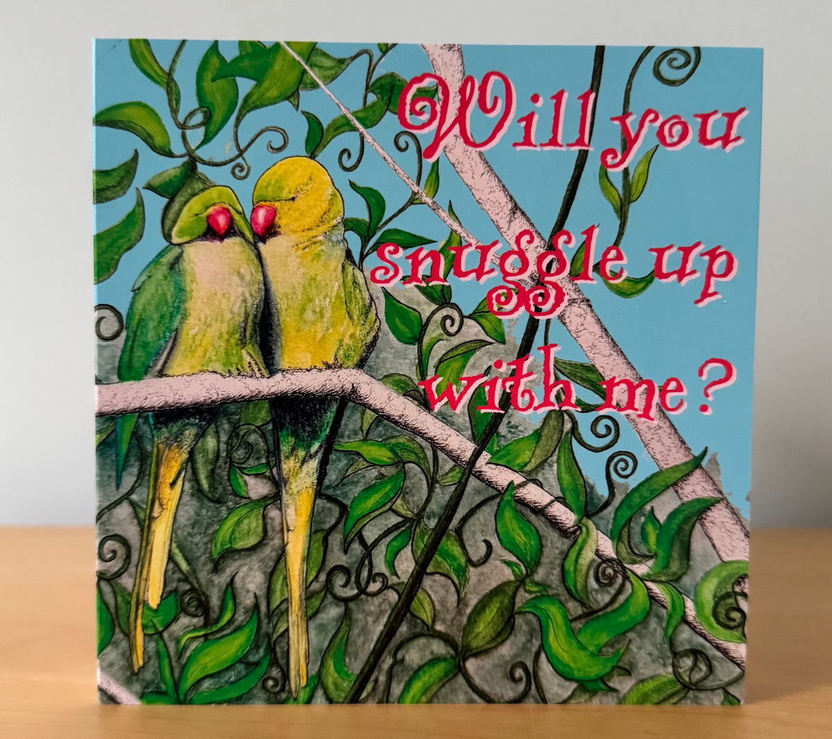 A square greeting card featuring an illustration of two green and yellow lovebirds on a branch, with red script text that reads 'Will you cuddle up with me?' against a light blue sky background. The card is standing on a light wood surface against a neutral gray wall.