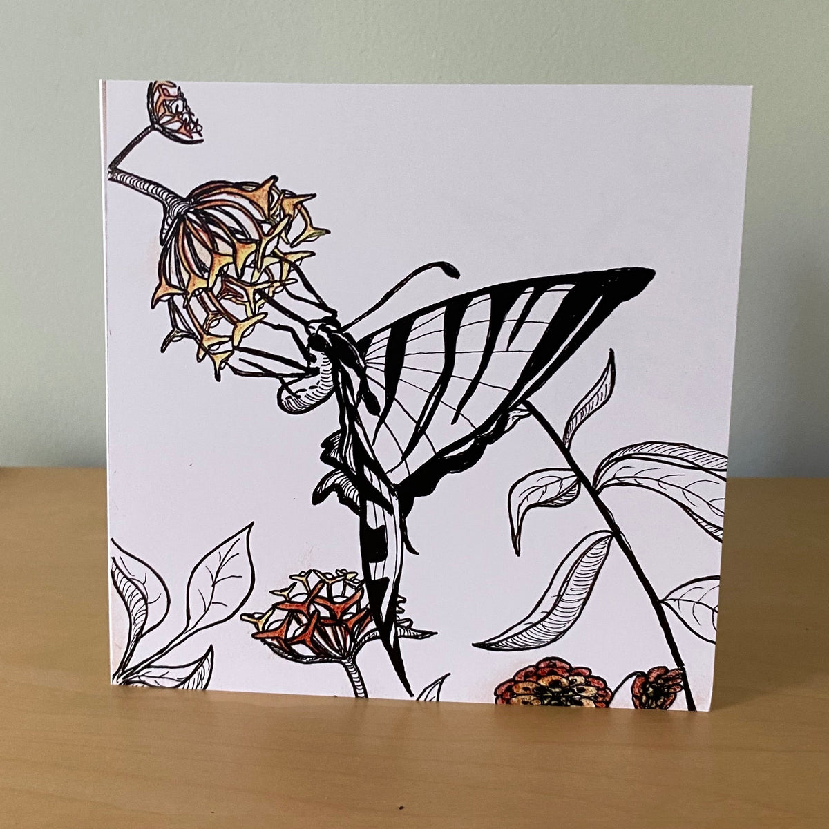 A square white art print or card featuring a black line drawing of a butterfly and flowers, standing on a light wood surface against a pale green wall.