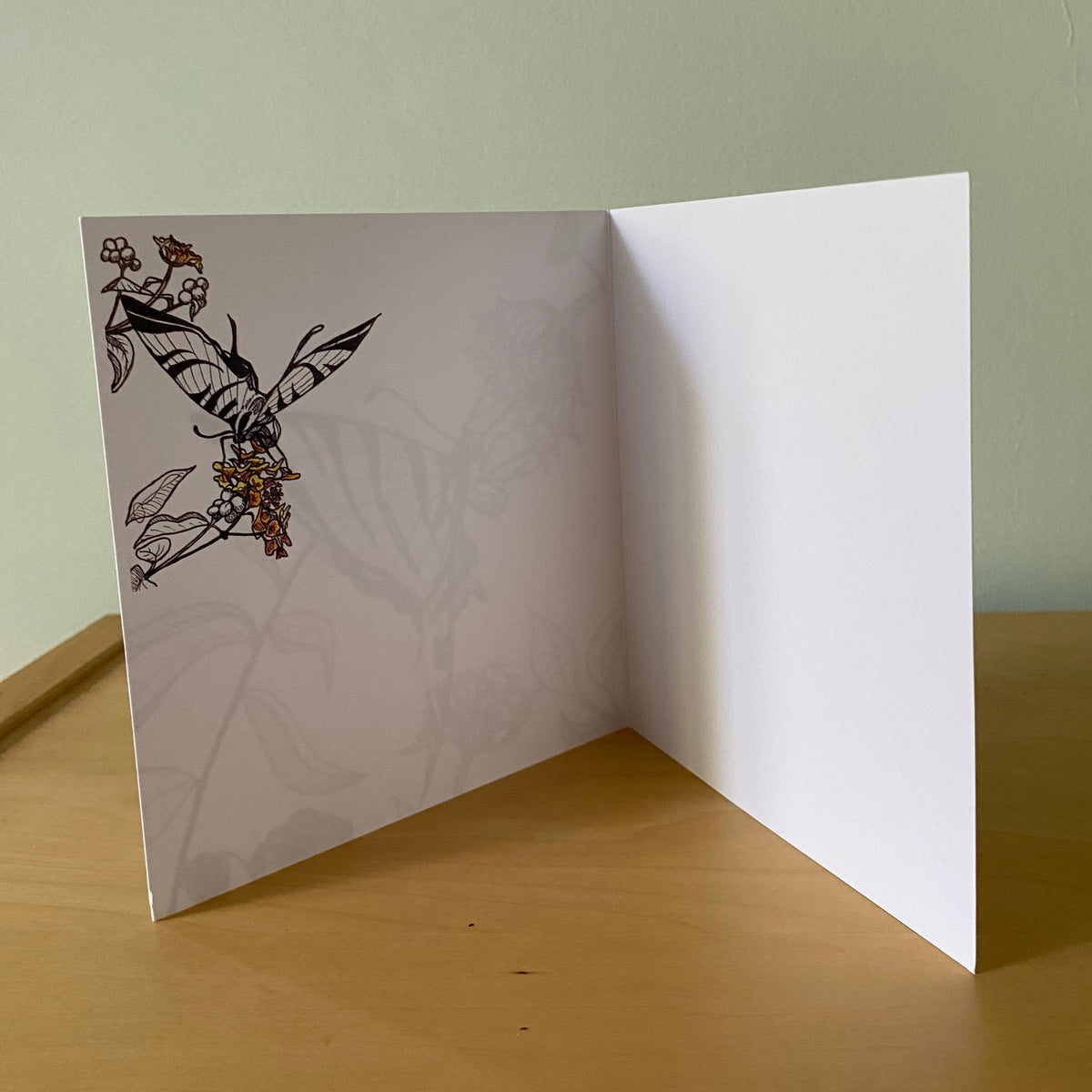 A white folded greeting card or art print standing open on a wooden table against a light green wall. The left panel features a black line-art illustration of a butterfly with small red and orange floral accents.