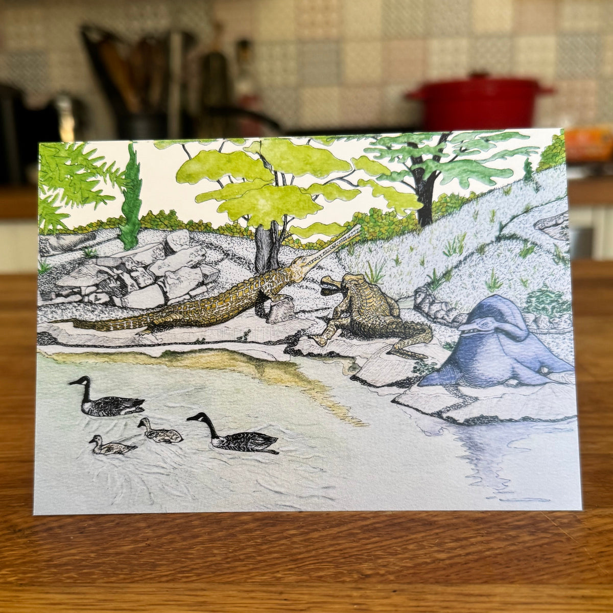 A square art print or card featuring a hand-drawn illustration of a river scene with dinosaurs, ducks, trees, and rocks, displayed on a wooden table with a blurred kitchen background.